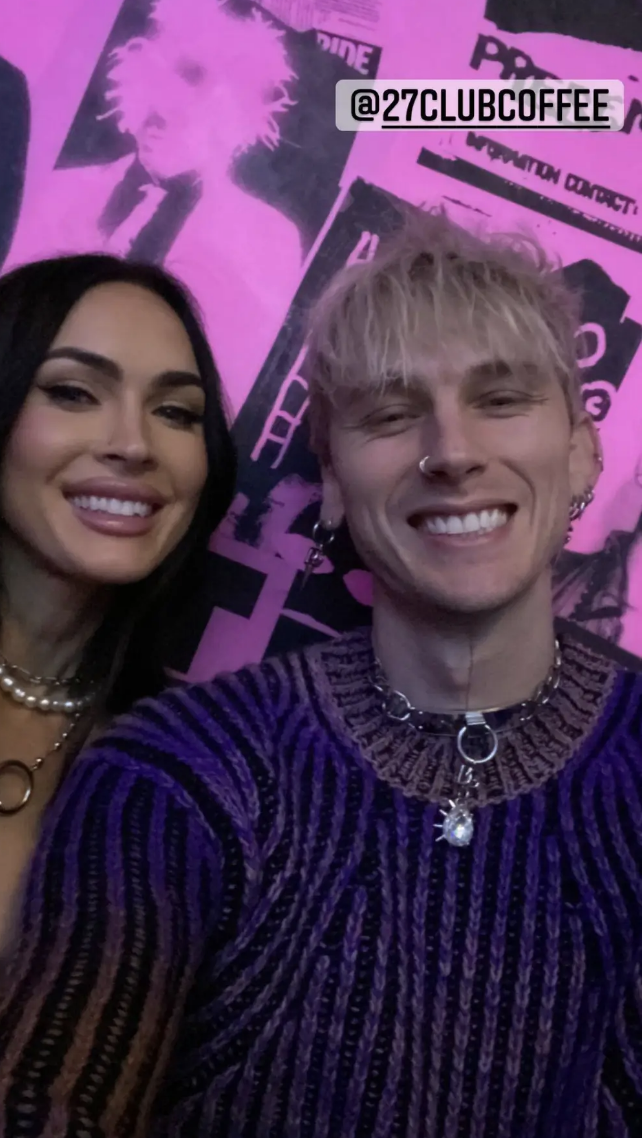 Megan Fox and MGK smiling closely for a selfie with a graphic poster in the background. MGK on the right wears a knit sweater