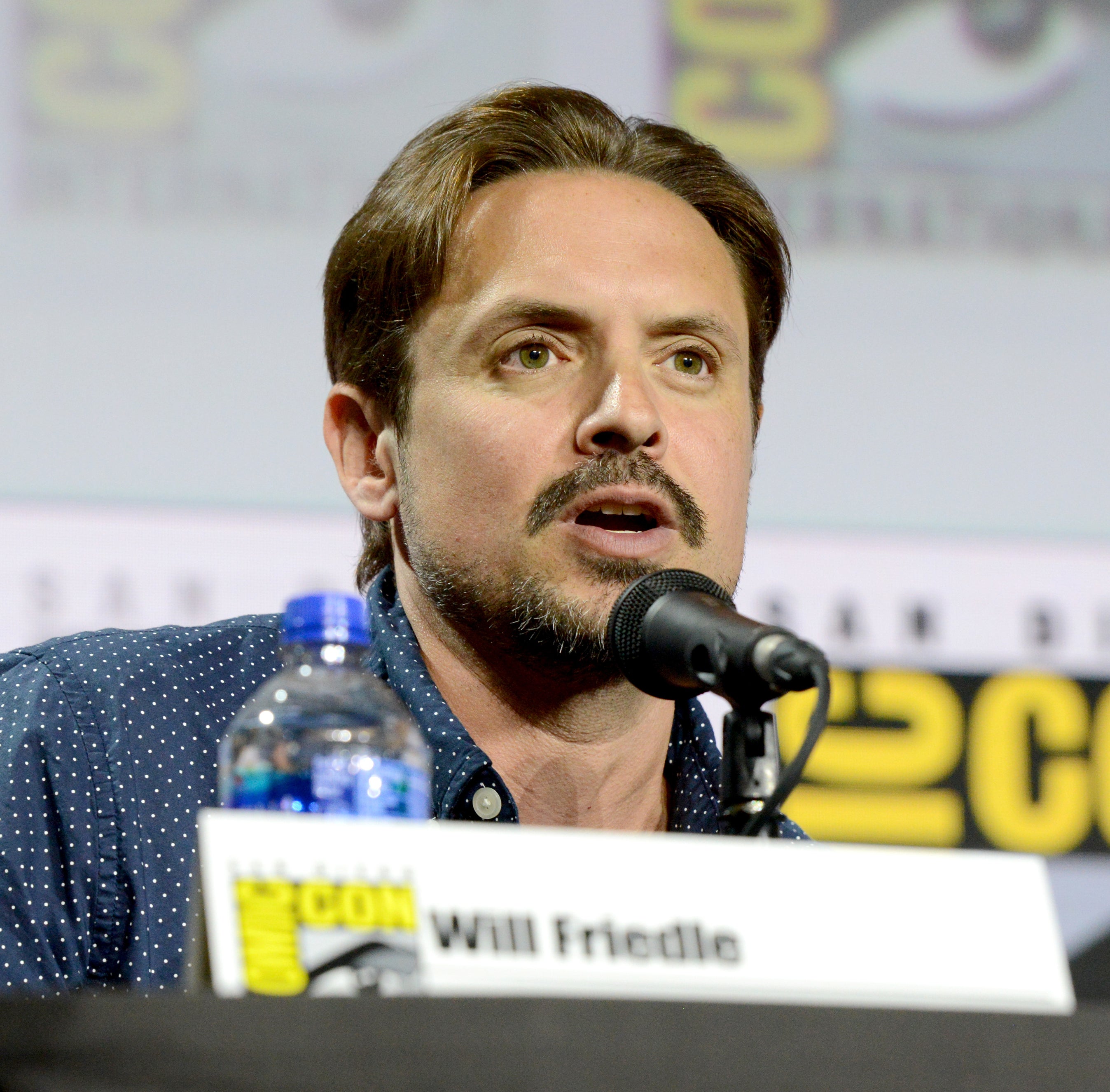 Will Friedle speaking at a Comic-Con panel, dressed in a button-up shirt, microphone in front