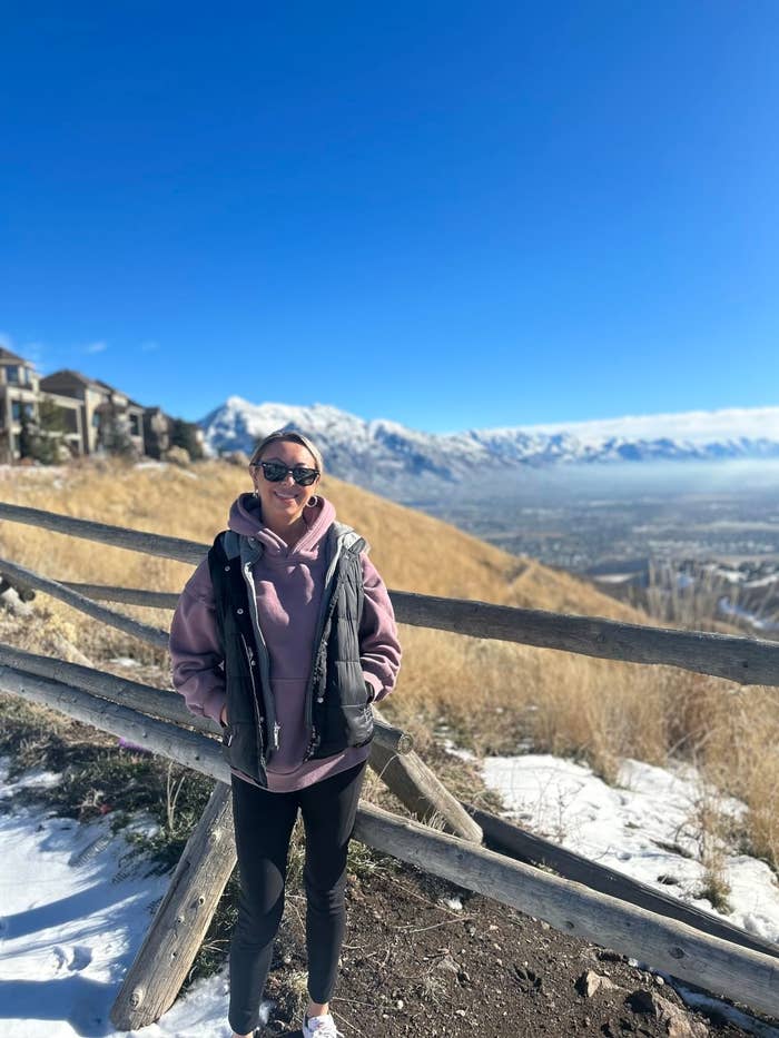 Person in a layered outfit with a jacket and sunglasses stands by a wooden fence with snowy mountains in the background