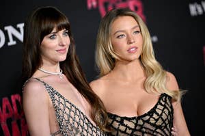 Two women in elegant dresses pose together at an event; one in a chainmail-style dress, the other in a lacy, scoop-neck gown