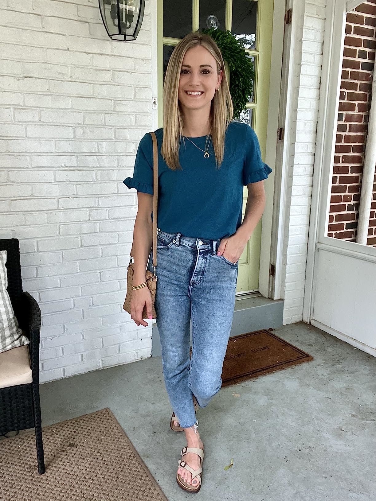 Reviewer in a casual chic outfit with a tucked-in tee, denim jeans, and strappy sandals standing on a porch