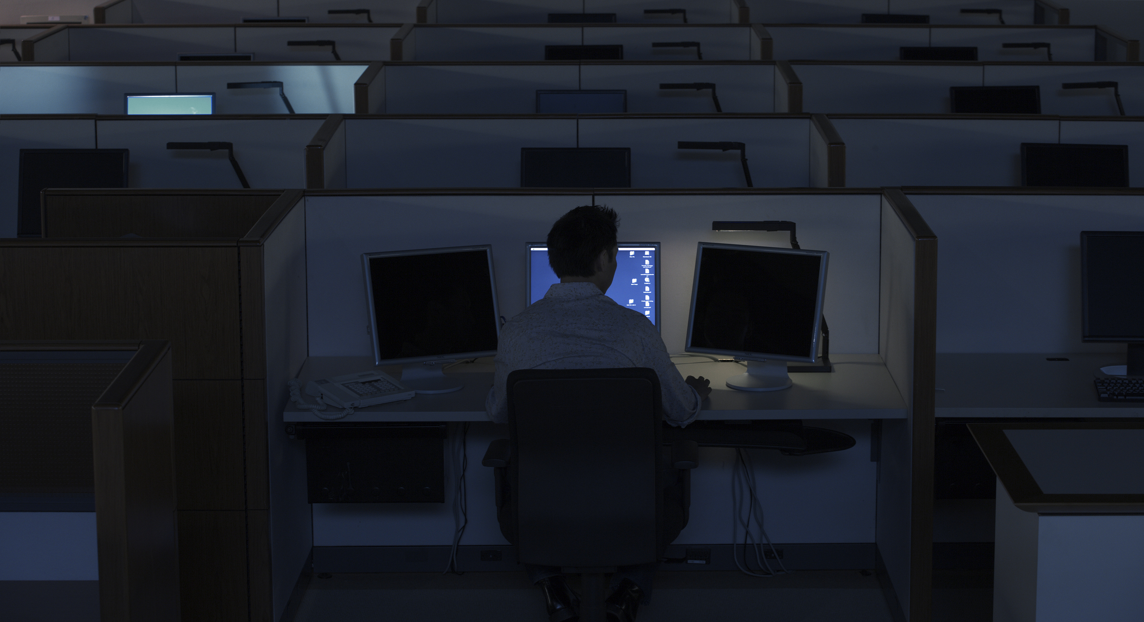 Person at a desk in a dark office with multiple computer screens, suggesting late-night work
