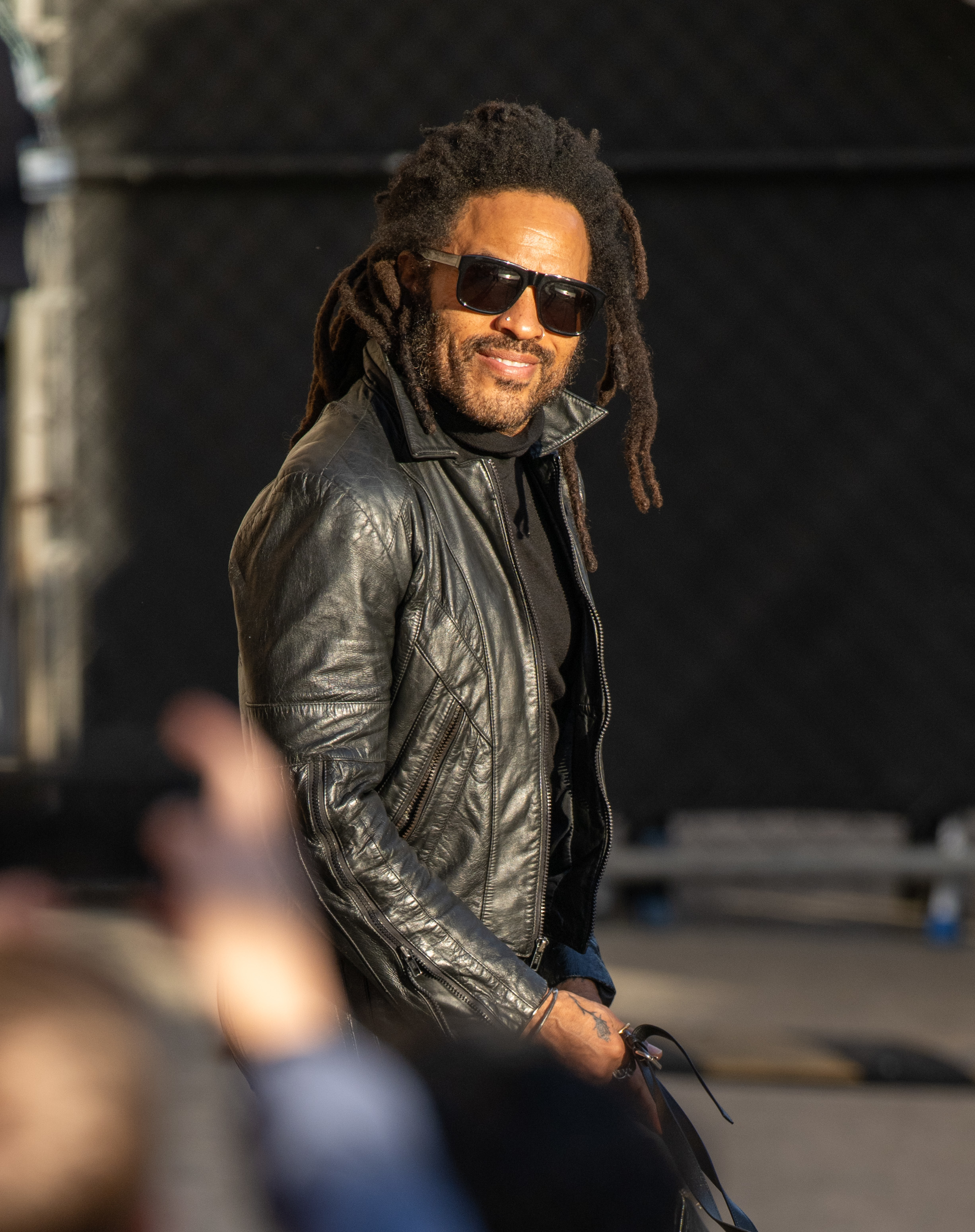 Lenny wearing a leather jacket and smiling as he walks past observers
