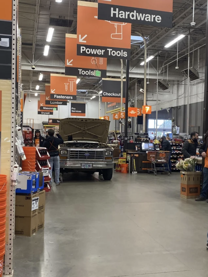 A vintage truck is displayed inside a hardware store with shoppers and aisles of products