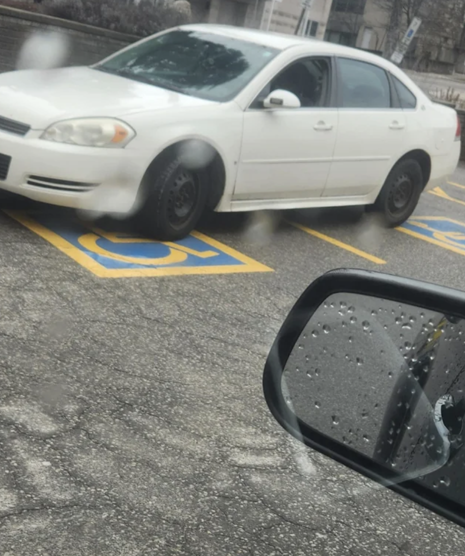 A white sedan parked diagonally across two accessible parking spaces with visible markings. Rearview mirror shows raindrops