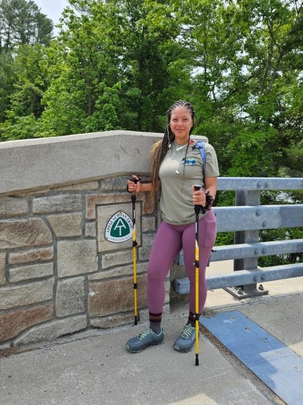 Reviewer with trekking poles stands by trail sign, wearing athletic leggings and top and a backpack