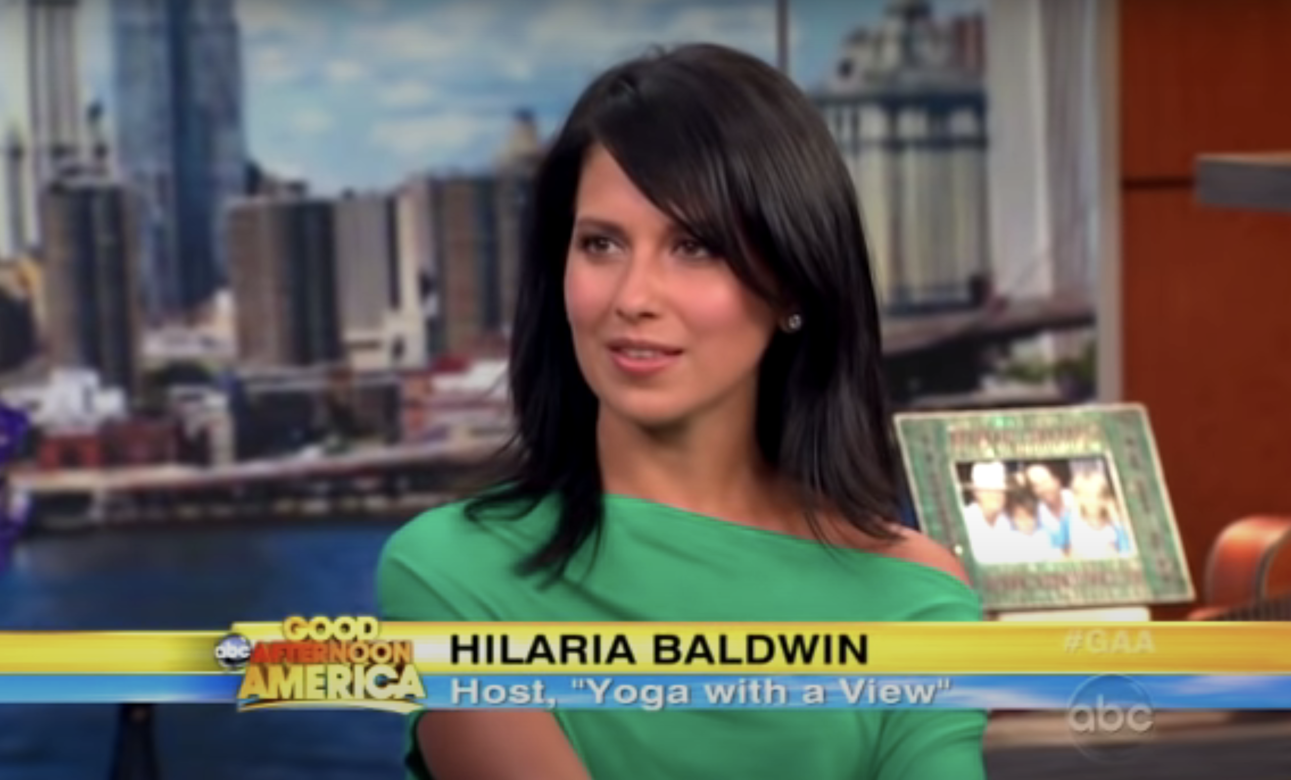 Hilaria Baldwin in a TV interview, wearing a solid green top with cityscape backdrop
