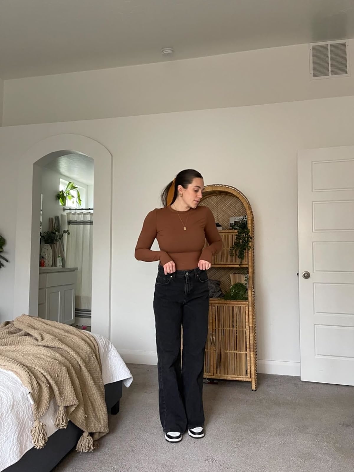 Individual in a fitted brown top and black jeans stands in a bedroom with minimal decor, near a rattan mirror