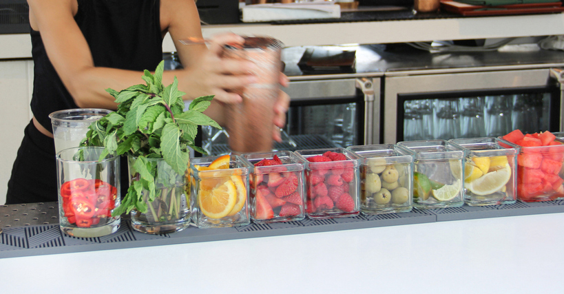 Person preparing a drink at a bar with various fruits and mint leaves displayed