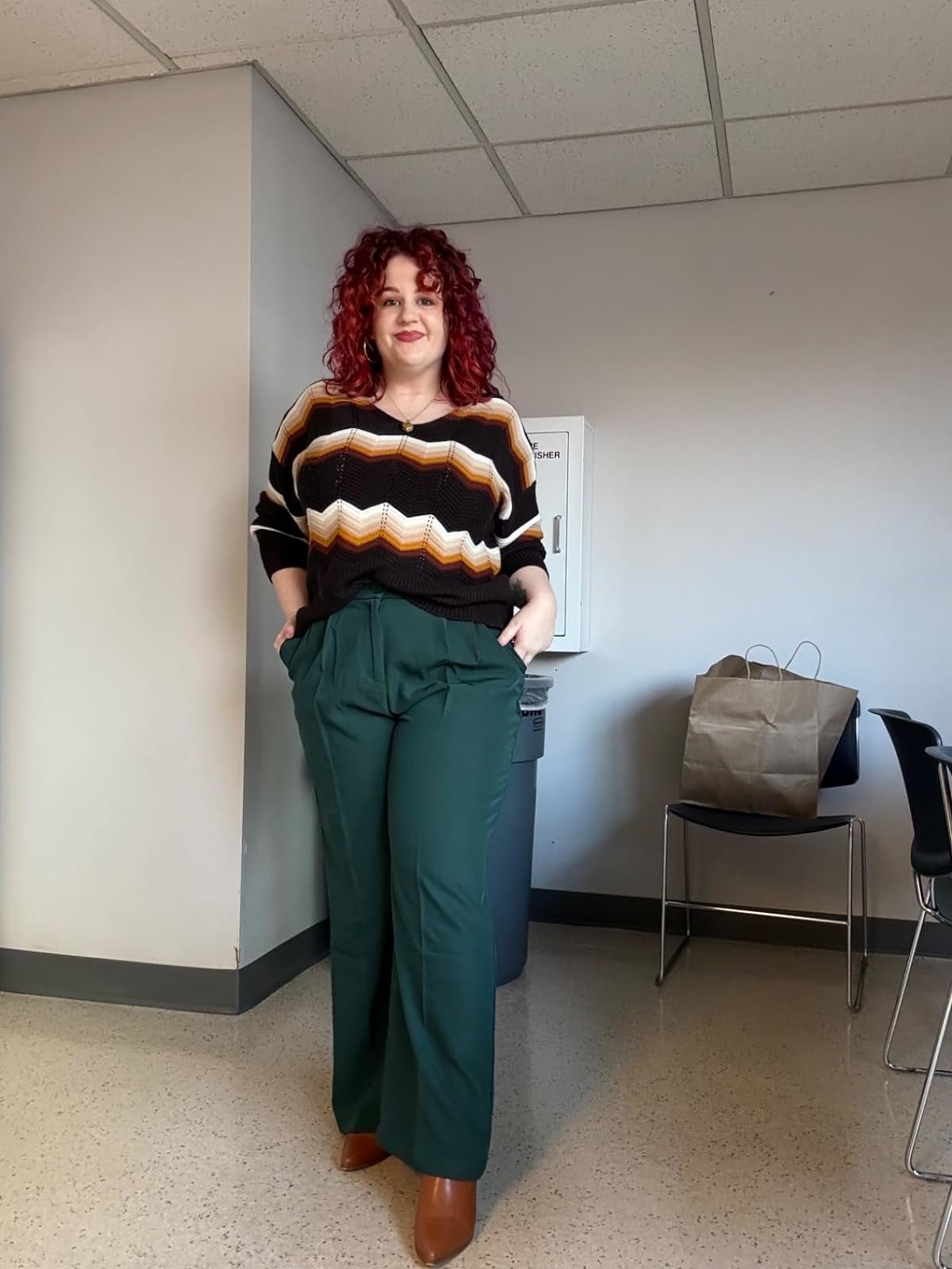 Model in a striped sweater and high-waisted green trousers, with hands in pockets, standing indoors