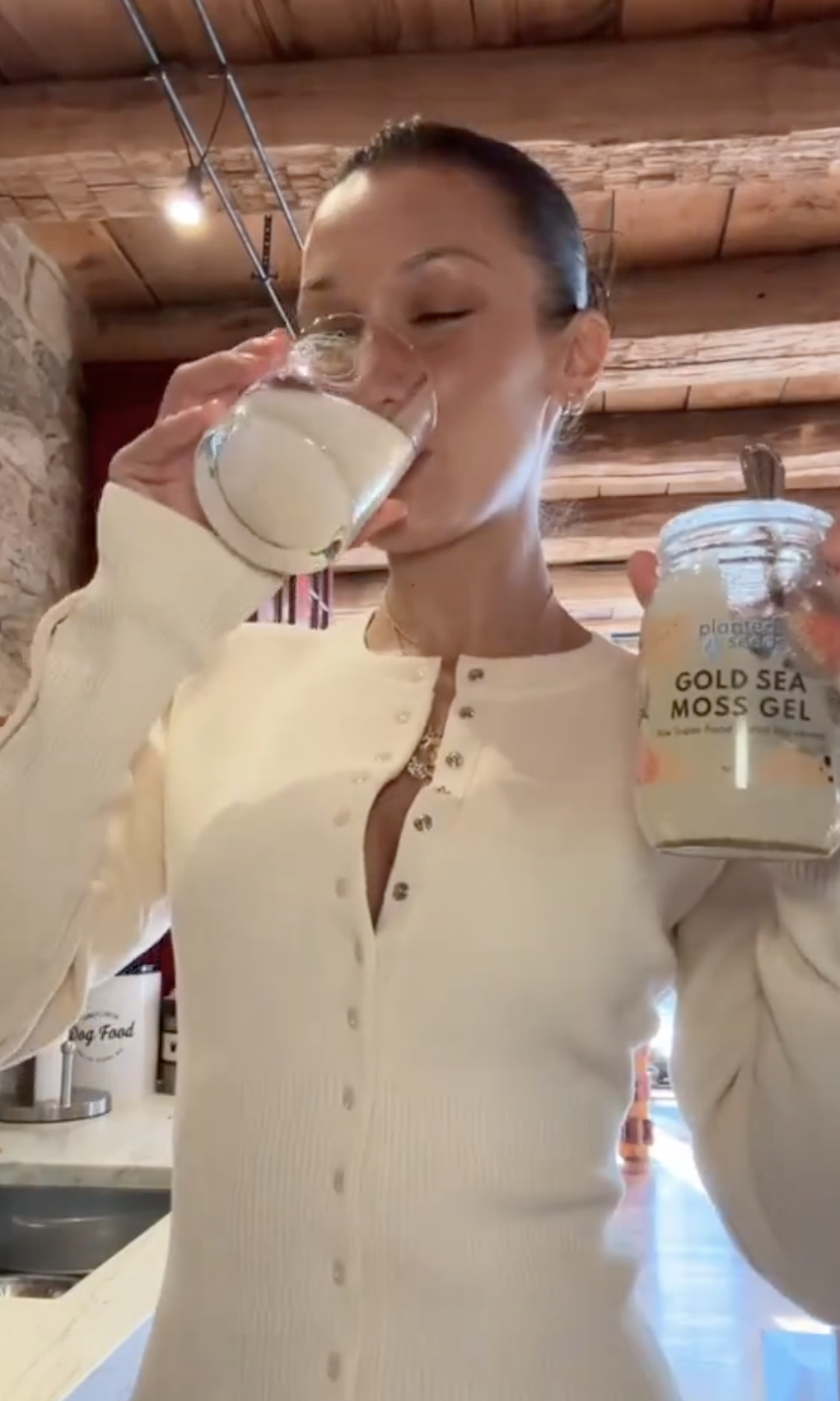 Bella in a buttoned top drinking a glass of a pale, cloudy beverage, holding a jar labeled &quot;Gold Sea Moss Gel&quot;