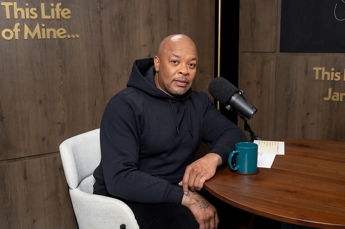 Dr. Dre sits at a podcast desk with a microphone and a mug. He wears a black hoodie