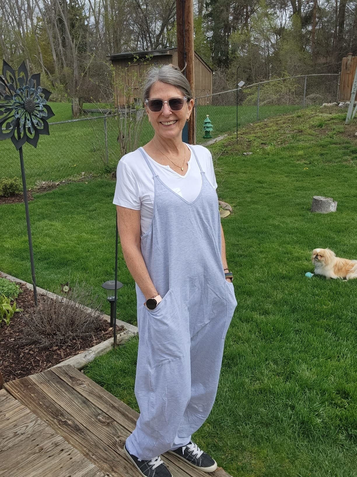 Model in casual jumpsuit with layered top, sneakers, standing in a garden with a dog in the background