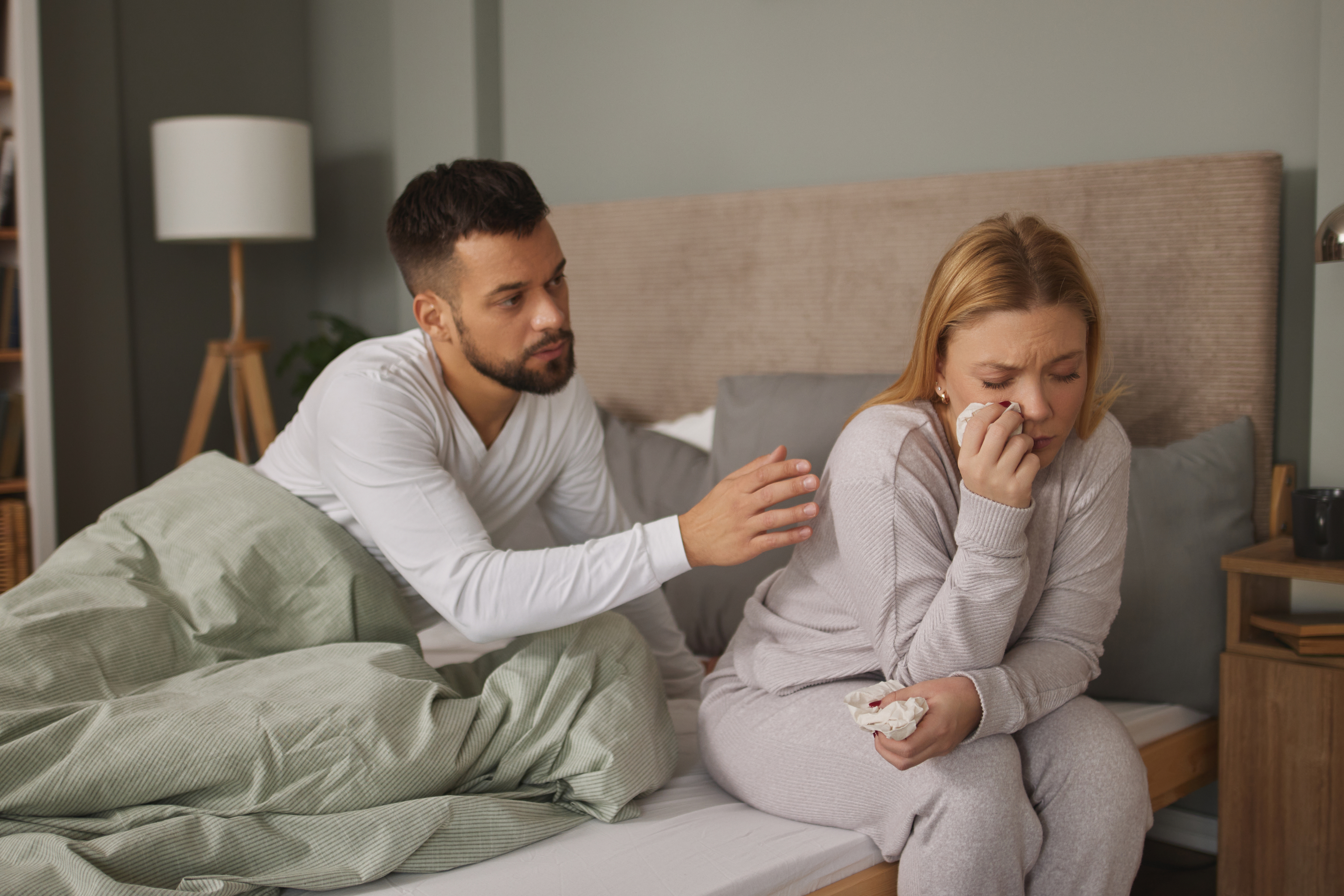 Man comforting upset woman sitting on bed, both in casual attire