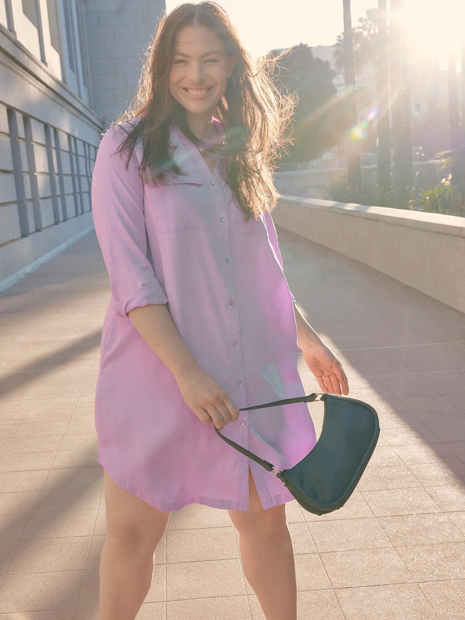 person in a buttoned shirt dress smiling with a sling bag, standing outdoors