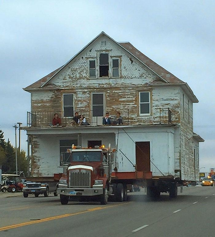 An old two-story house is being transported on a large trailer with people sitting on the porch roof on the highway