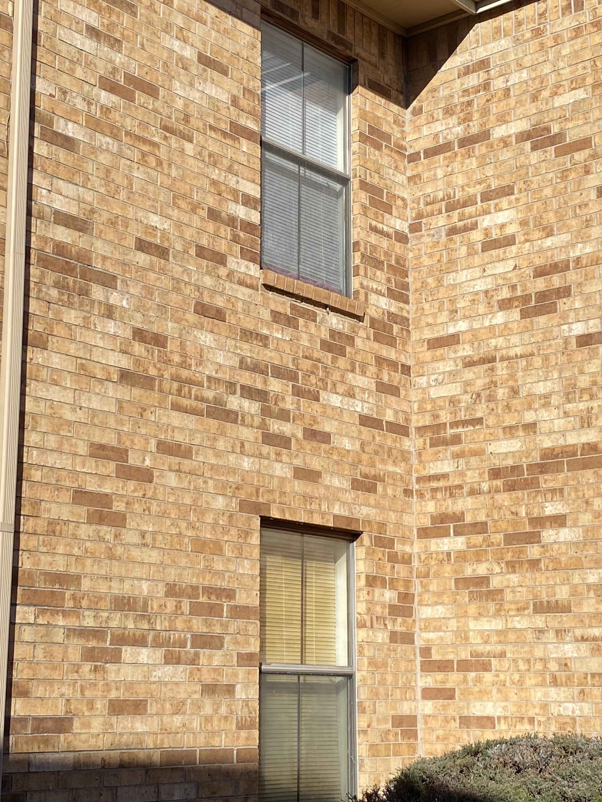 Two stacked windows with blinds on a brick wall, with the blinds of one window light brown and other white