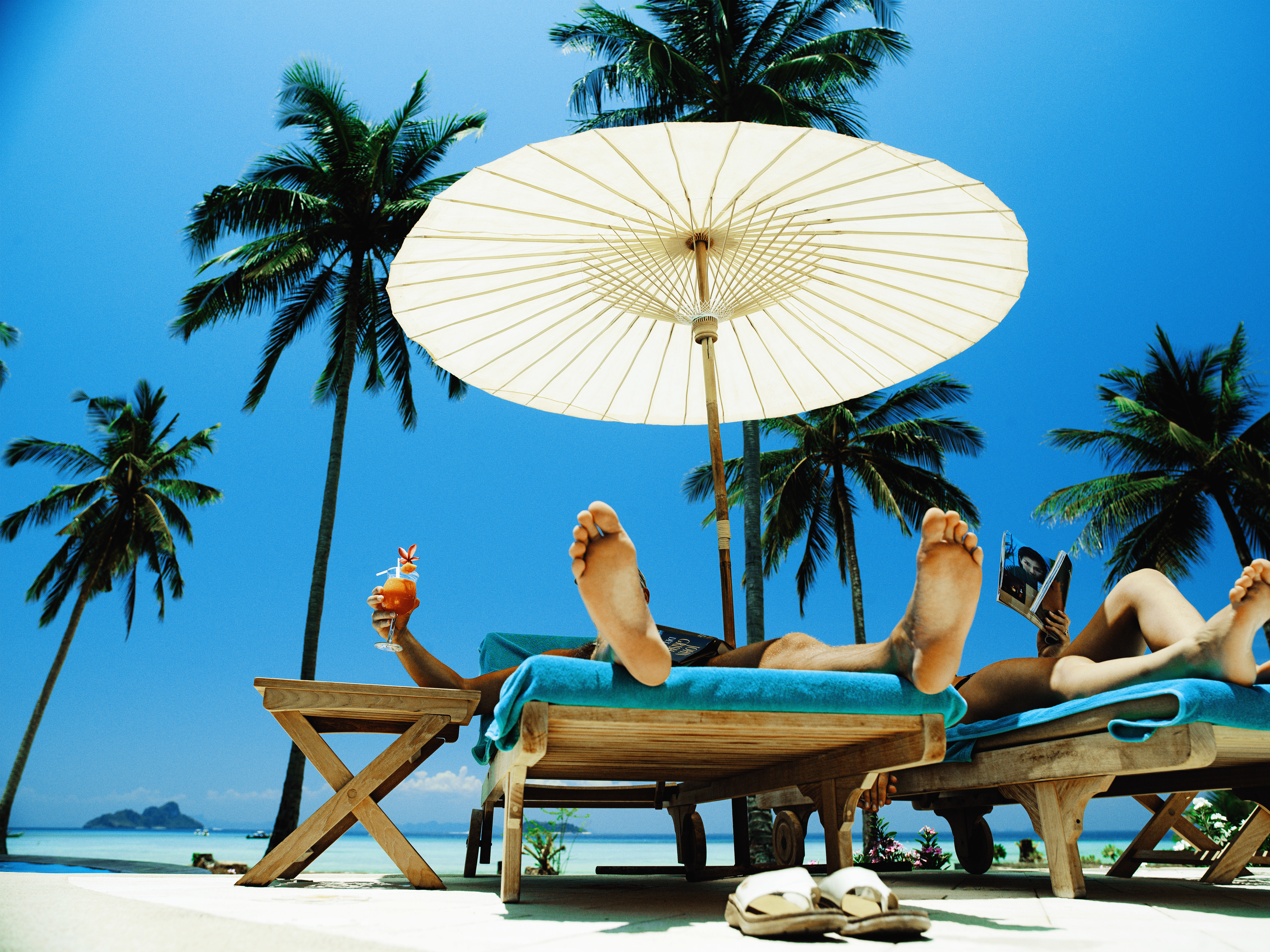 Two people relaxing on sun loungers under a large umbrella, tropical beach setting