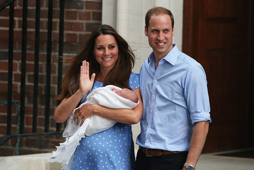 Kate Middleton in polka-dot dress holding a newborn, with Prince William in a button down shirt, both smiling