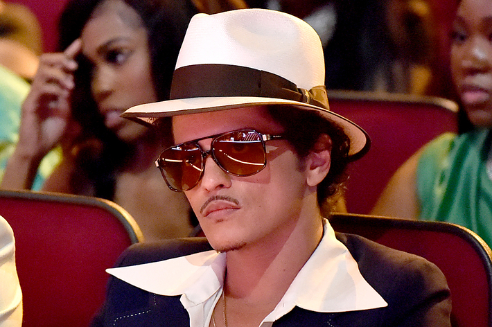 Bruno Mars at a music event wearing a white fedora, sunglasses, and a black suit with a white shirt