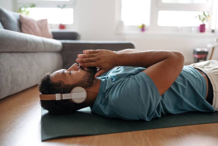 Man lying on a yoga mat indoors with headphones, resting with one hand on forehead