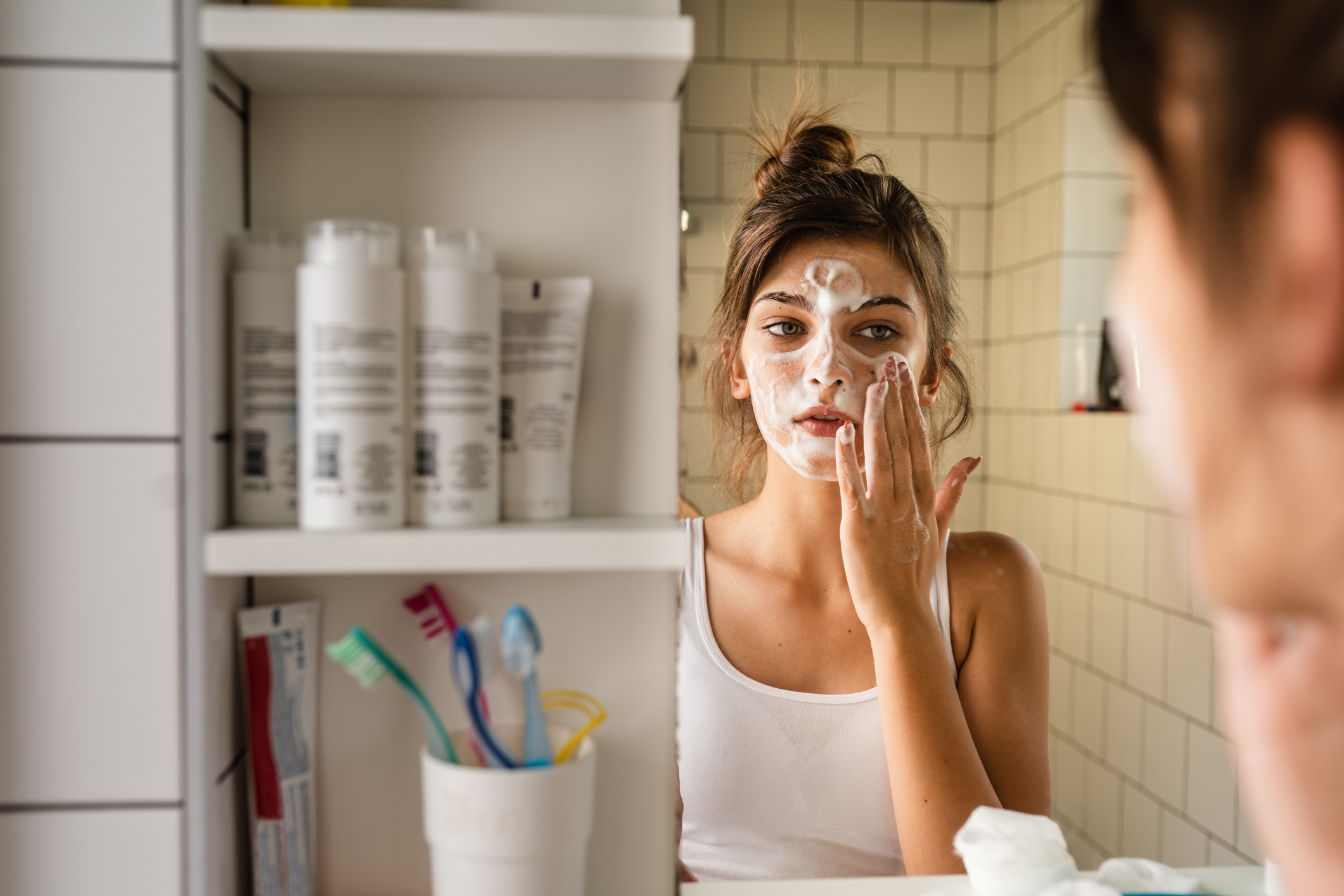 girl in front of mirror applies facial cream, with open cabinet and skincare products in background
