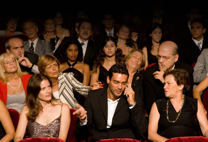 A man in a suit on the phone in a theater while everyone angrily looks at him