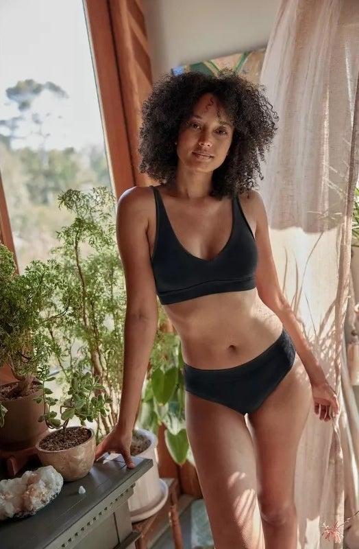 model in a casual black bralette and matching underwear set stands beside indoor plants
