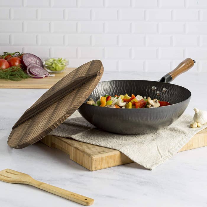 Wok on a kitchen counter with a wooden lid, containing sautéed vegetables, next to ingredients and utensils