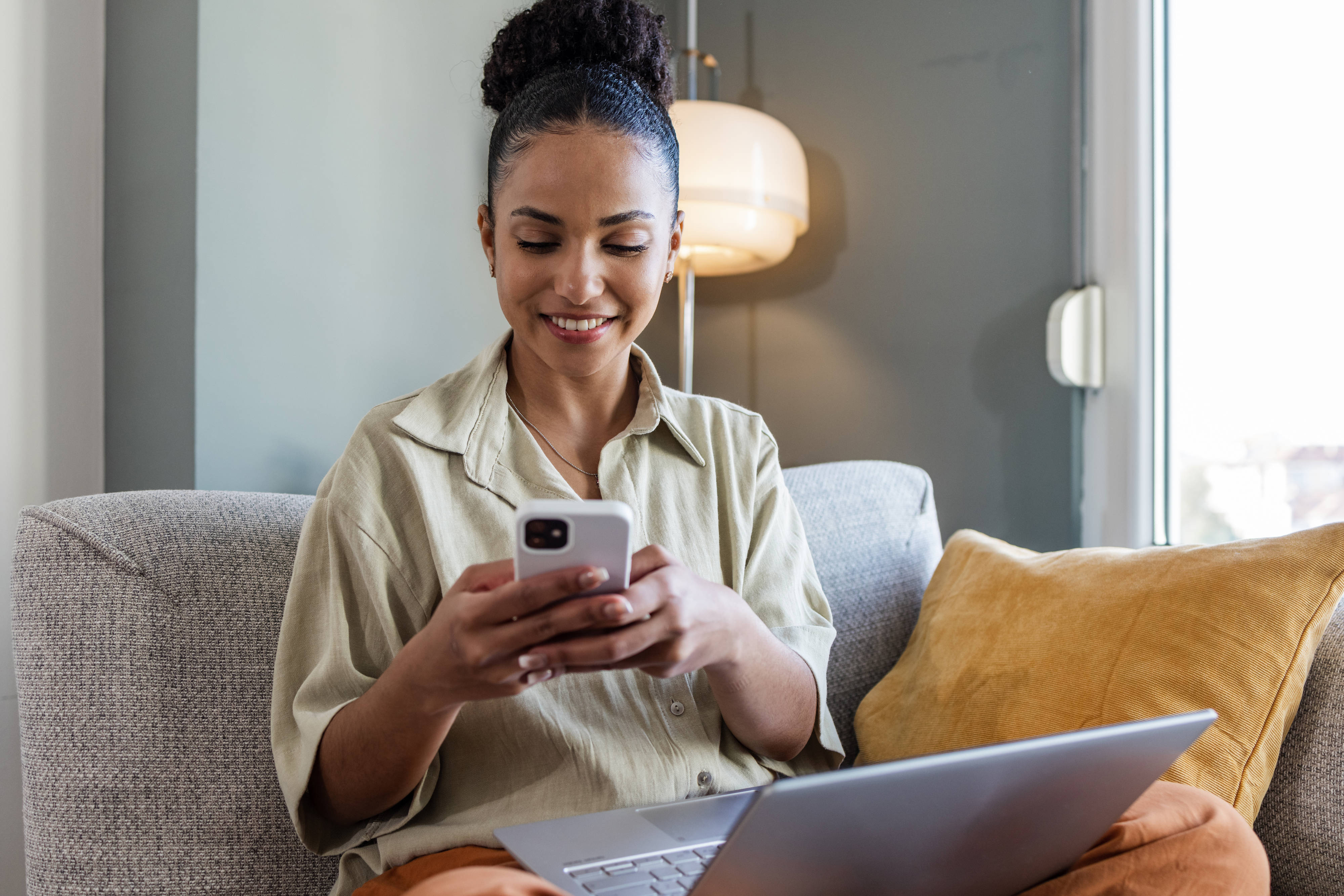 Woman on sofa with laptop, smiling at smartphone in her hands