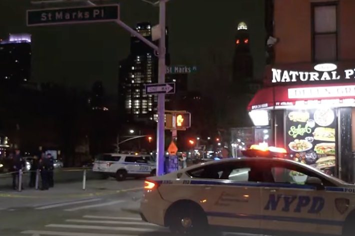 NYPD car in front of Natural Plus Deli on a street corner at night, city lights and buildings in the background