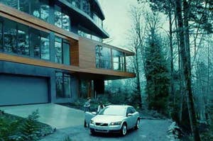 Modern house in a forest with a silver car parked outside. Two people are standing near the car