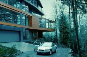 Modern house in a forest with a silver car parked outside. Two people are standing near the car