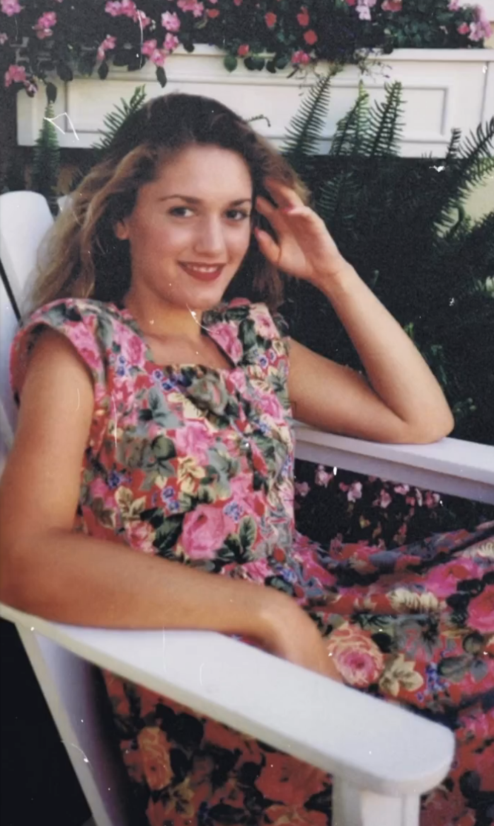 Gwen seated in a floral dress, smiling and touching her hair, in front of foliage