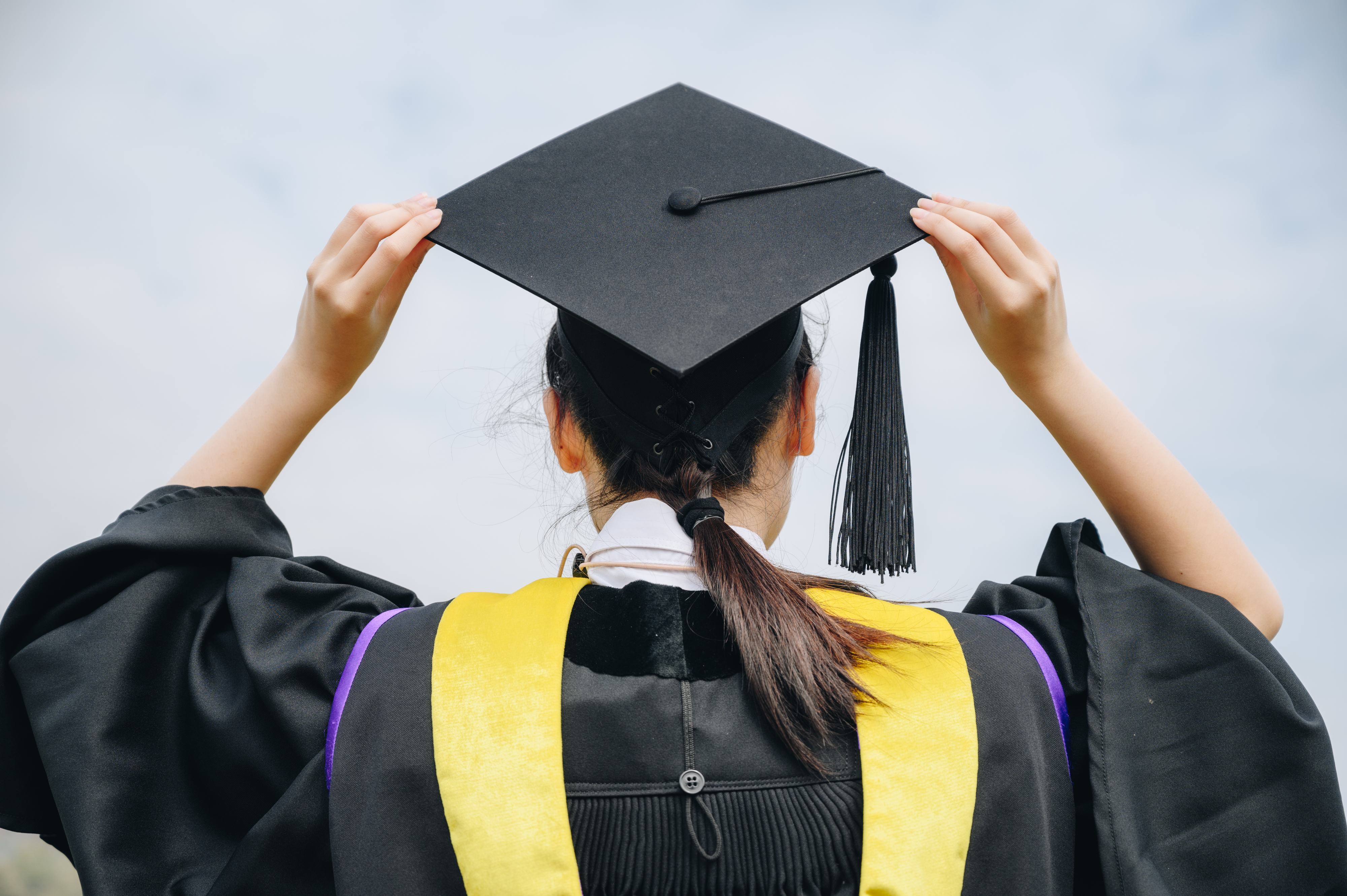 Graduate holding up a cap, seen from behind, wearing a gown with a yellow stole, symbolizing academic achievement