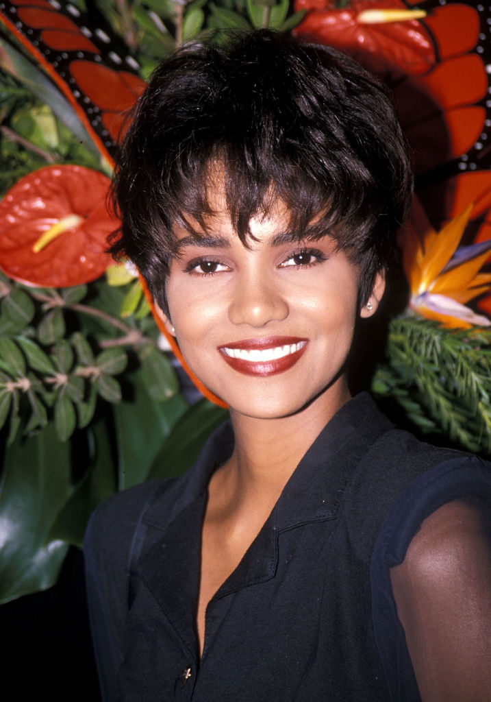 Halle with short hair, wearing a dark blazer, in front of tropical flowers