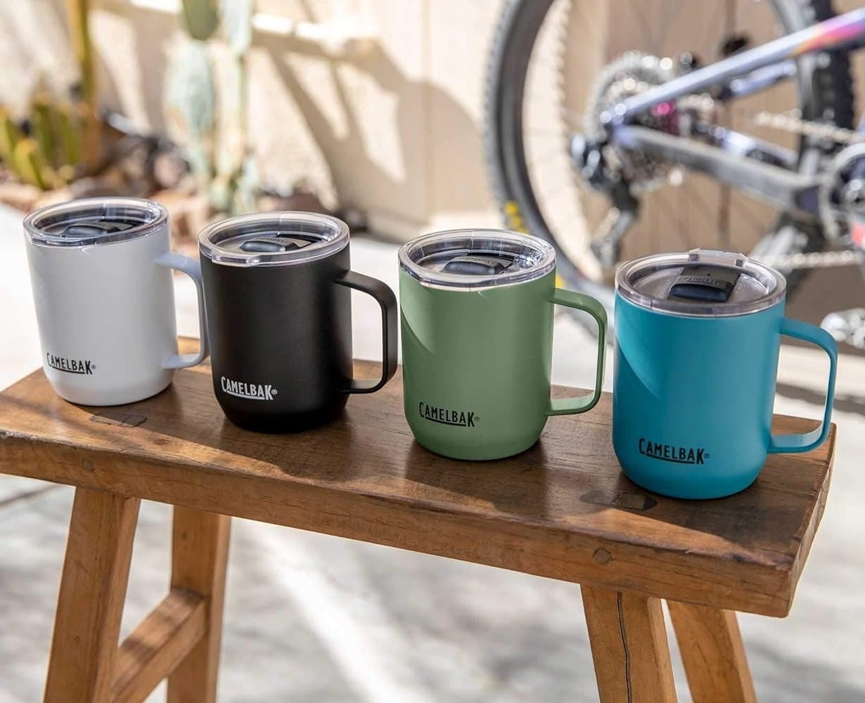 Four CamelBak travel mugs in white, black, green and blue on a wooden stool outdoors, with a bike in the background
