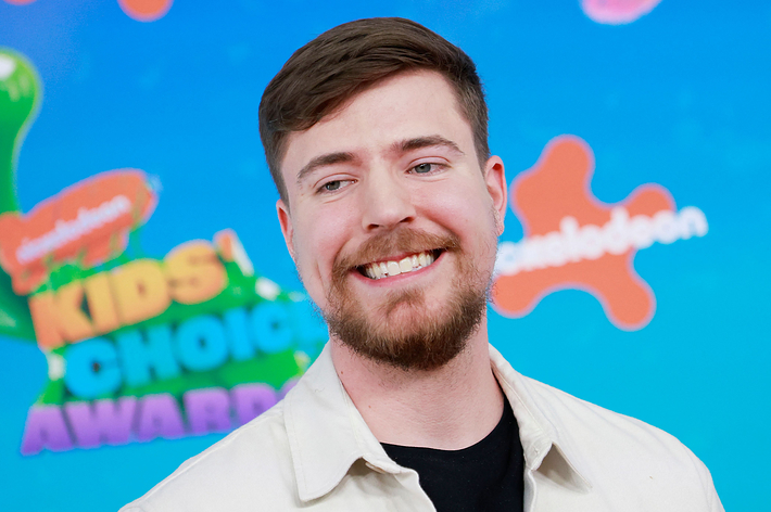 Smiling man at the Kids' Choice Awards with a logo backdrop, wearing a casual beige jacket