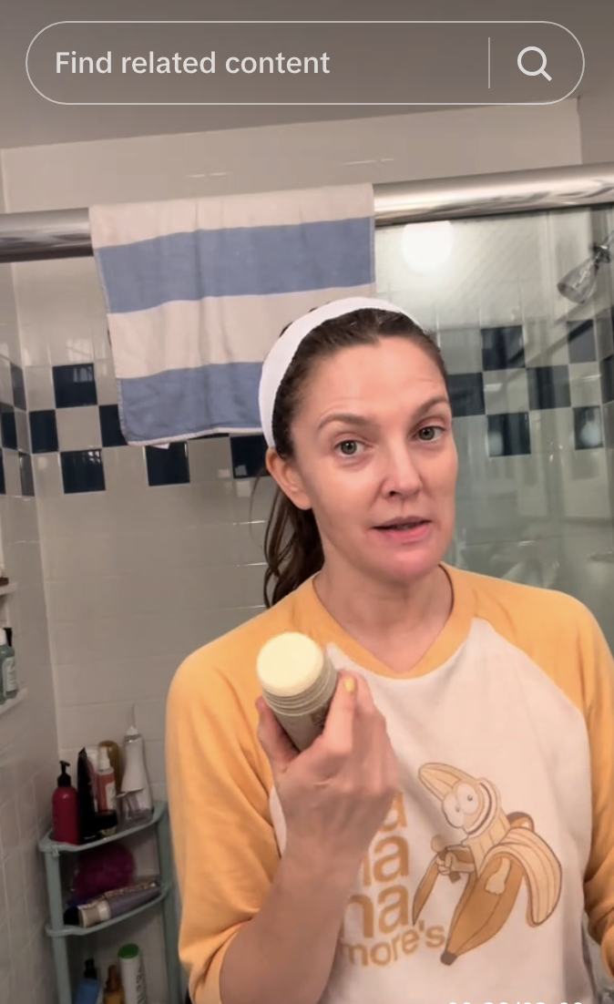 Drew Barrymore in bathroom holding a skincare product, wearing a headband and casual shirt with a banana graphic