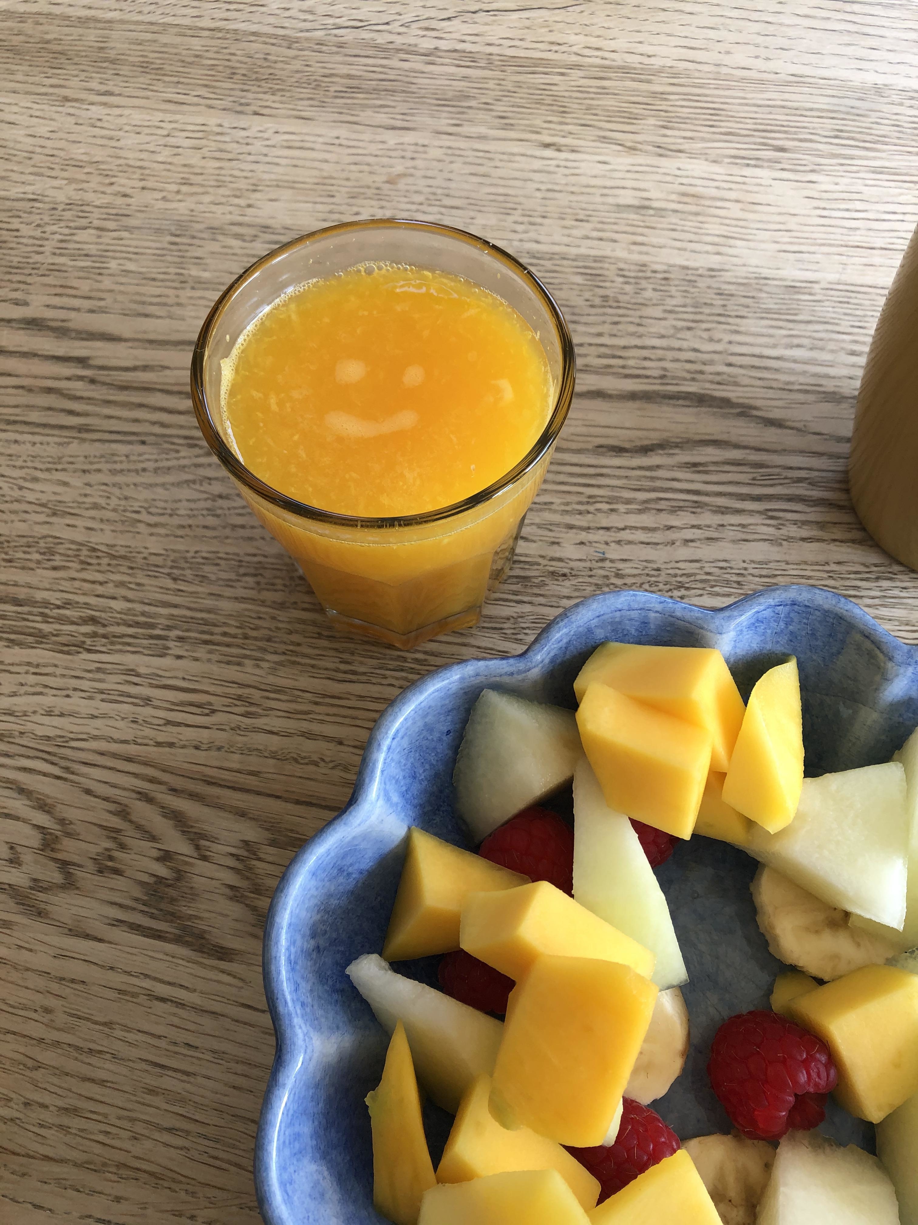 Glass of orange juice next to a plate of mixed fruit including mango, apple, and raspberries