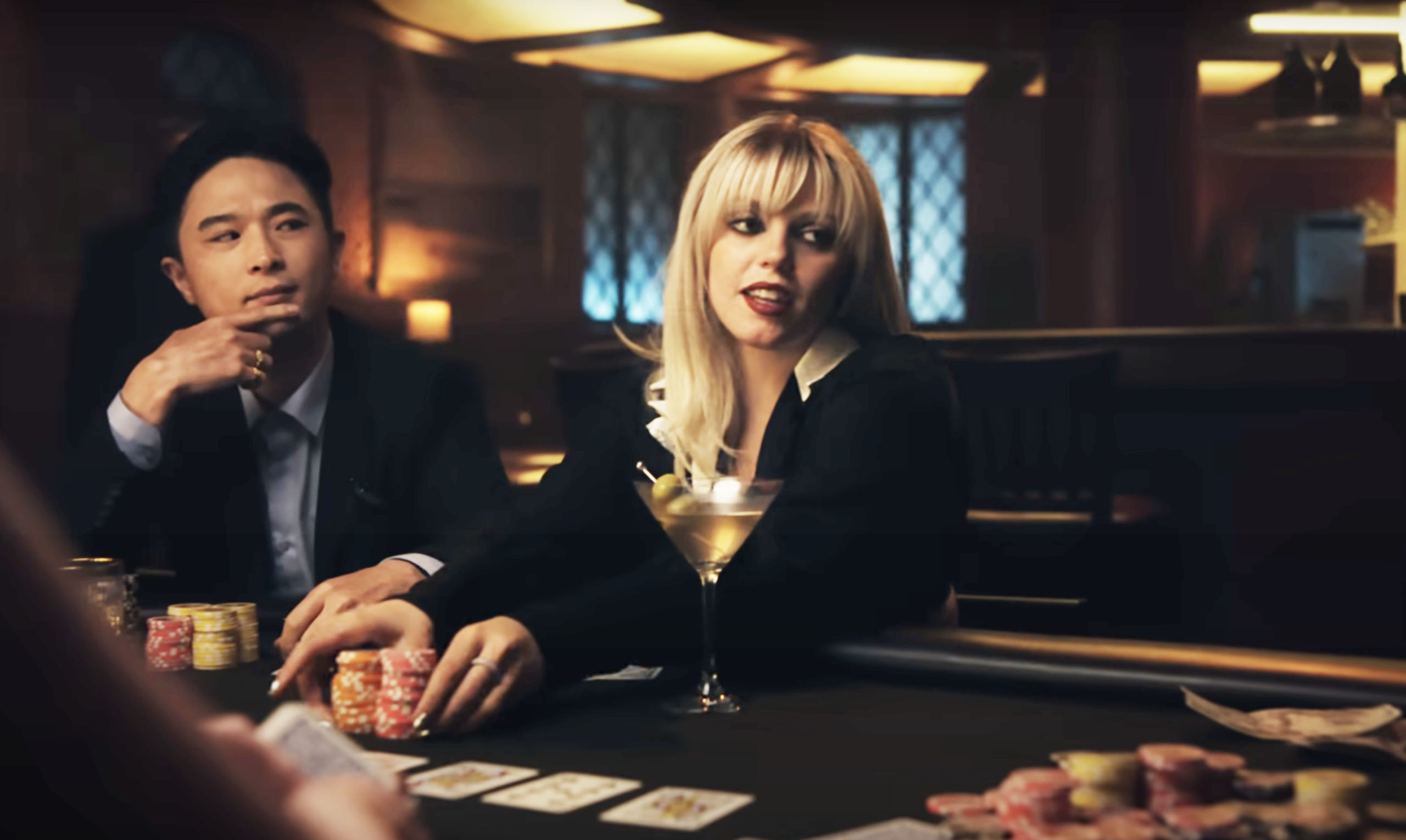 Two actors at a poker table in a scene, one with chips in hand, the other gesturing, with cards and chips on the table