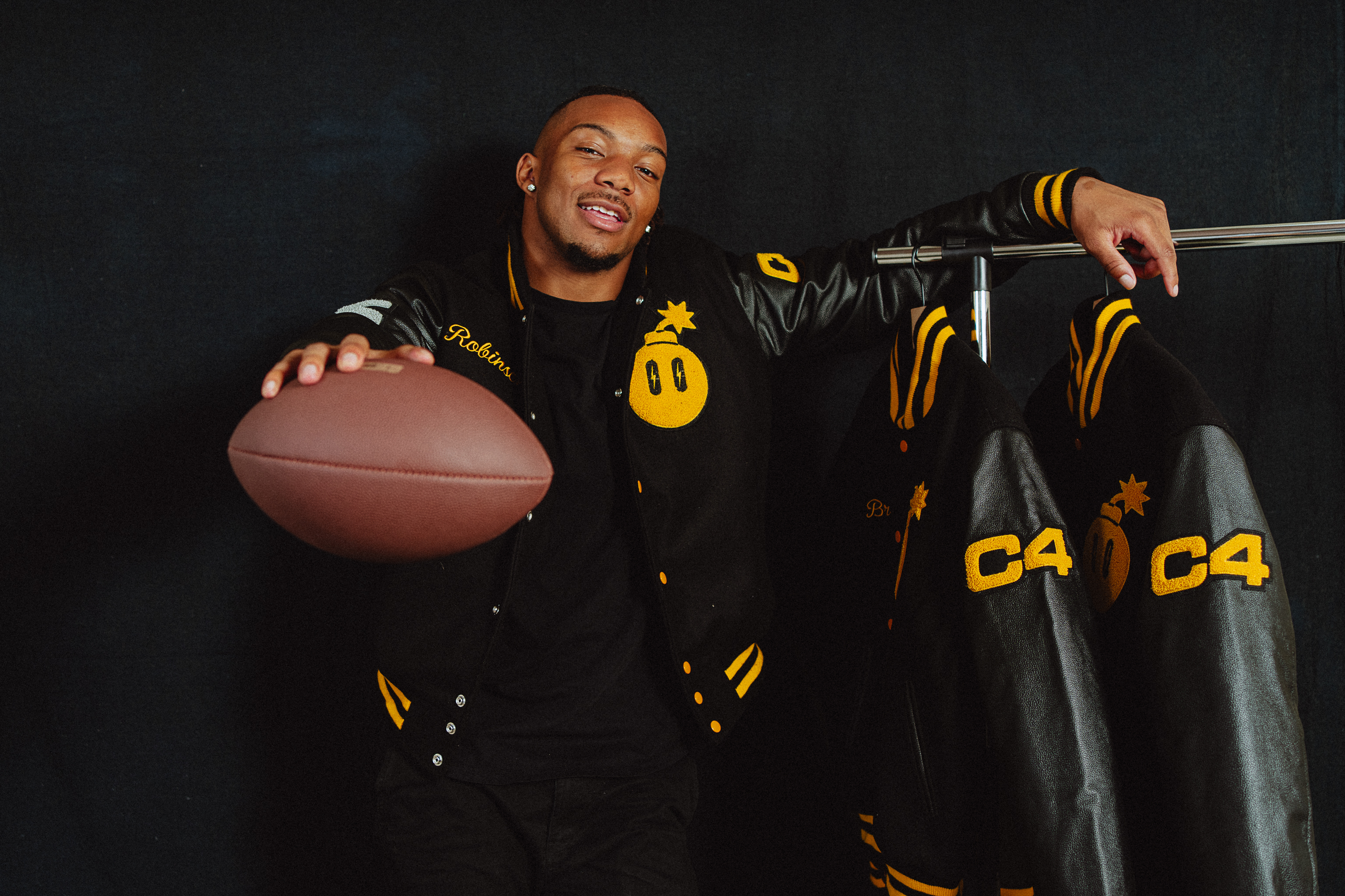 Man posing with football, wearing black and yellow sports jacket with smiley faces, leaning on rack with similar clothing