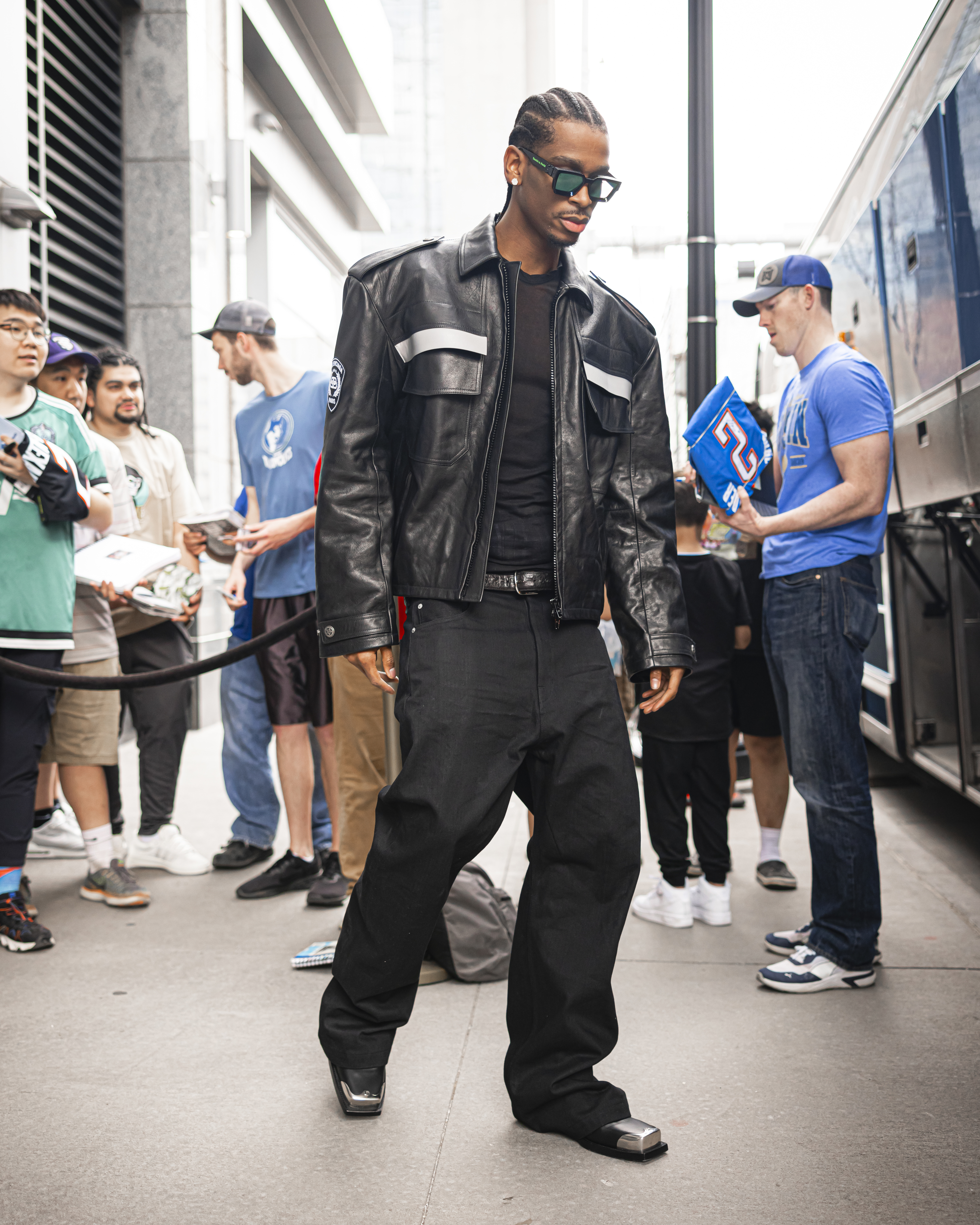 Person in a black leather jacket and pants walks past bystanders, possibly a sports figure arriving at an event