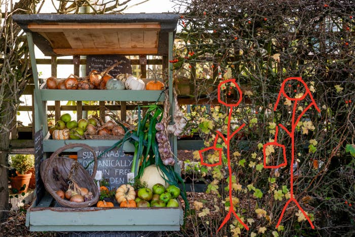 Wooden stand with a variety of fresh produce and a sign promoting sustainable gardening, along with two stick figures, each holding a bag