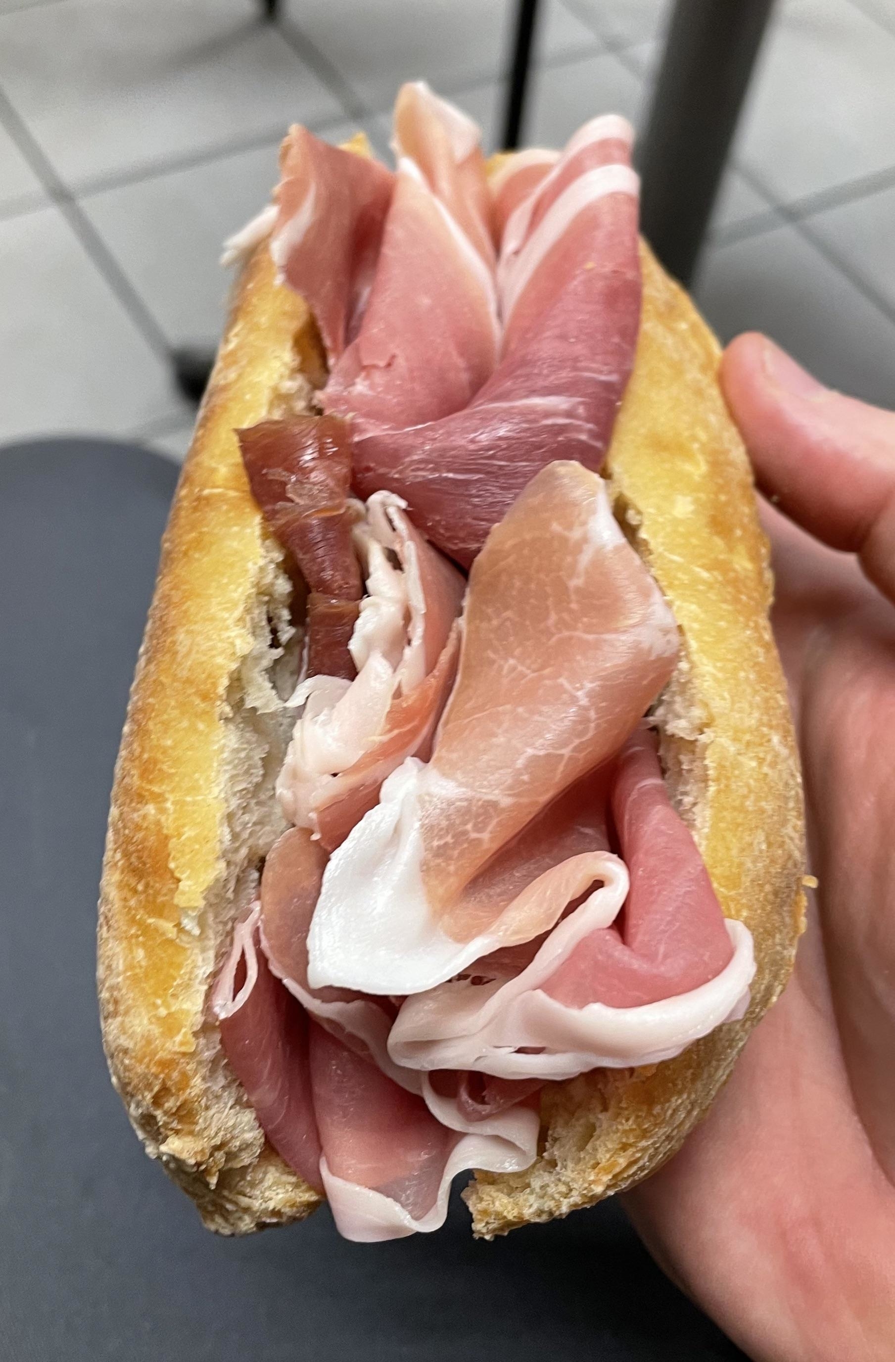 A hand holding a baguette sandwich filled with slices of cured ham