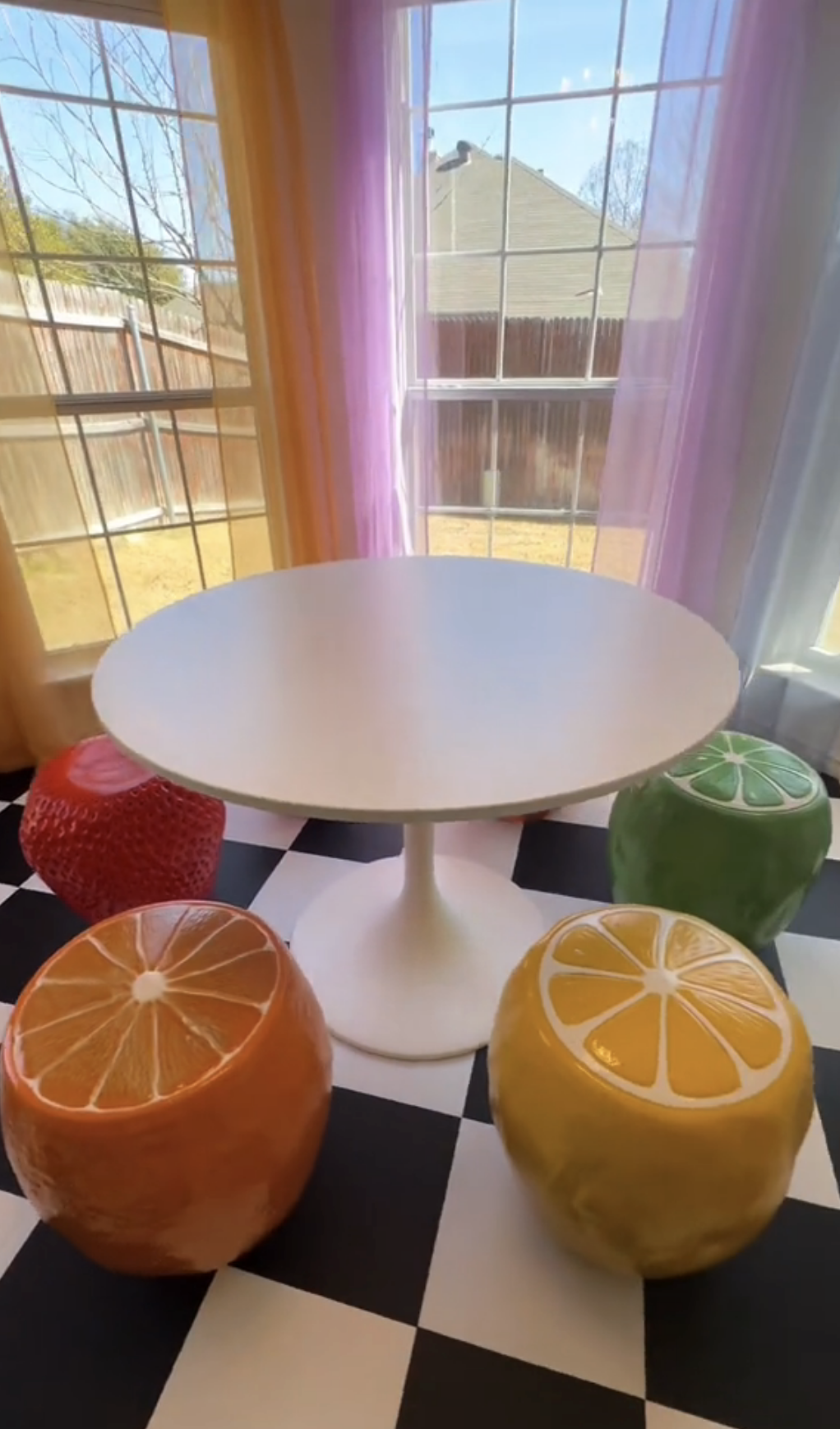 Table with fruit-shaped stool chairs on a checkered floor by a window with purple curtains