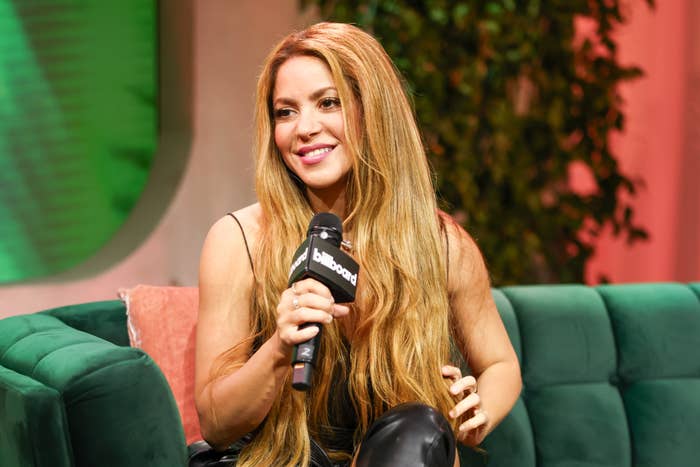 Shakira seated, holding a microphone, with a smile