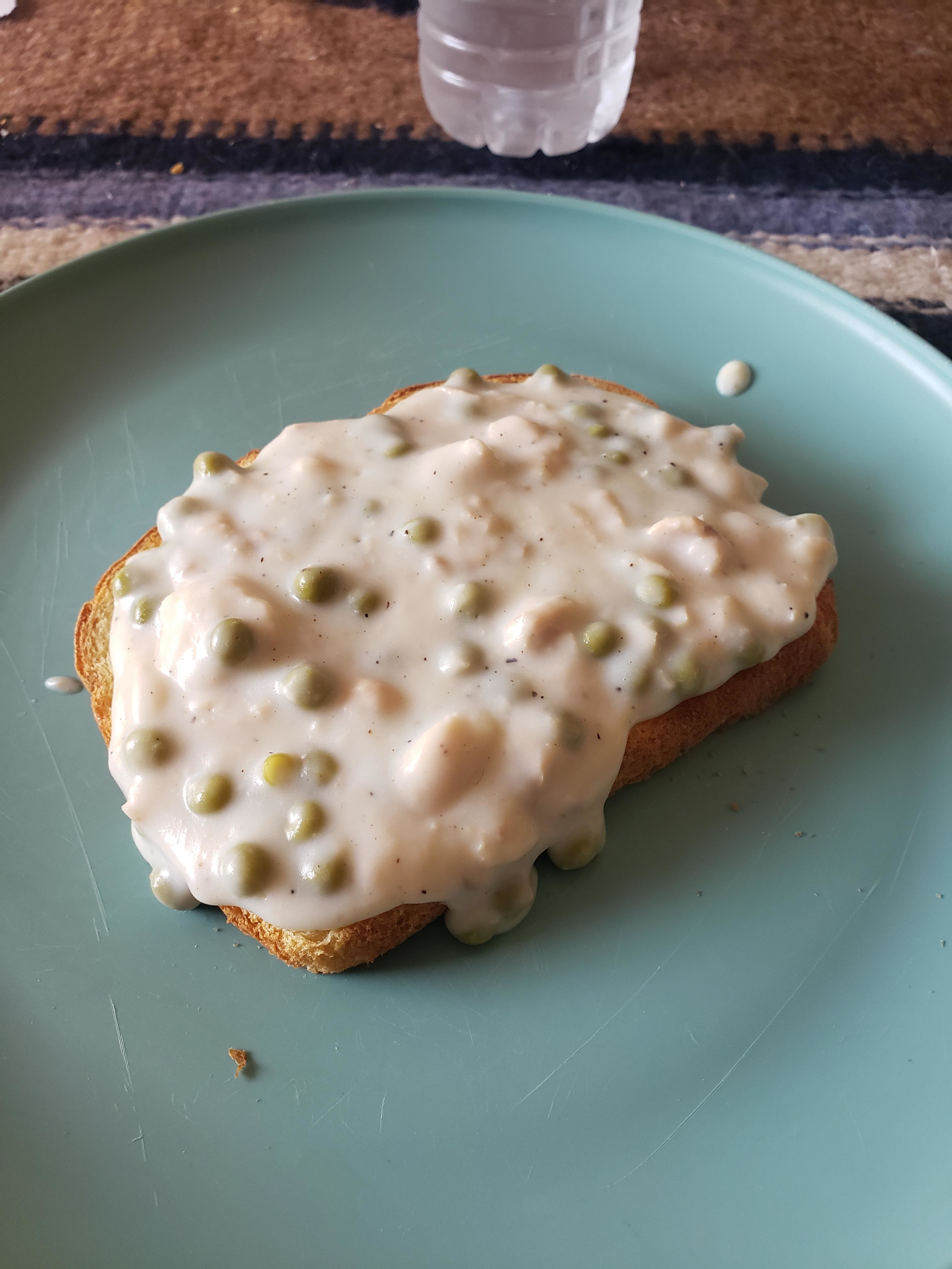 A piece of toast on a plate, topped with a creamy sauce with green peas