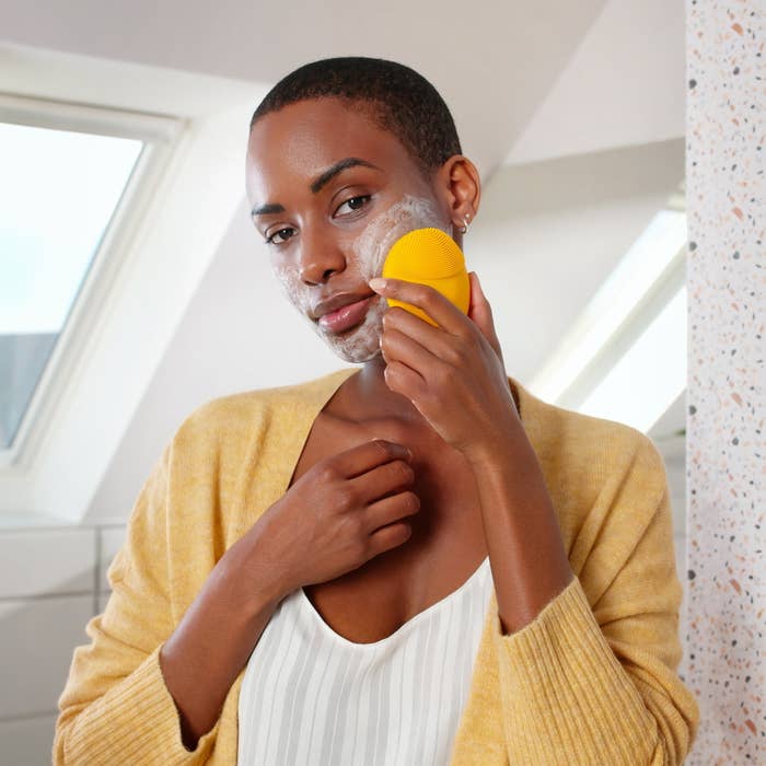 Person using a facial cleansing brush on their cheek, wearing a casual sweater