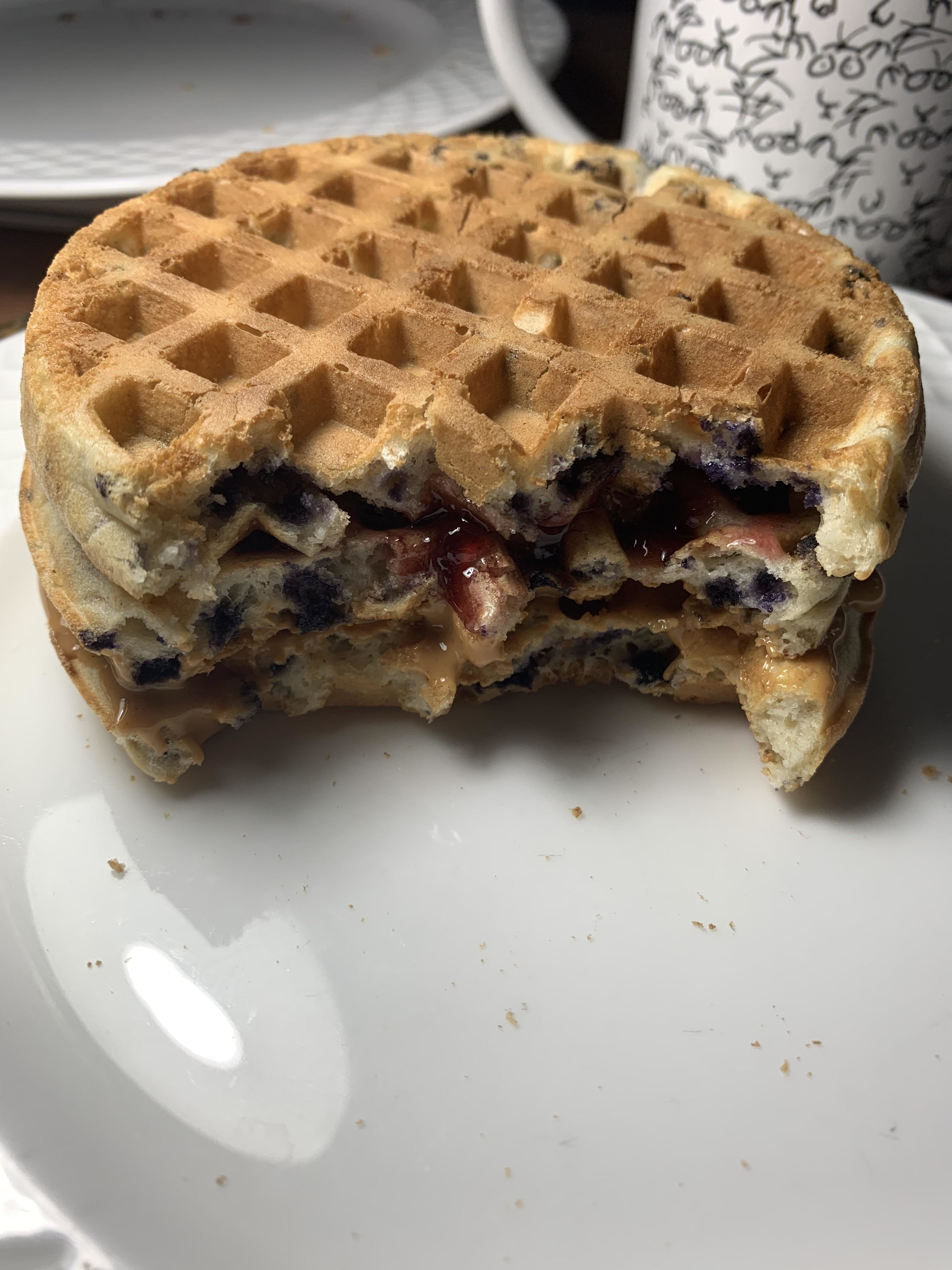 A half-eaten blueberry waffle sandwich with peanut butter and jelly on a white plate